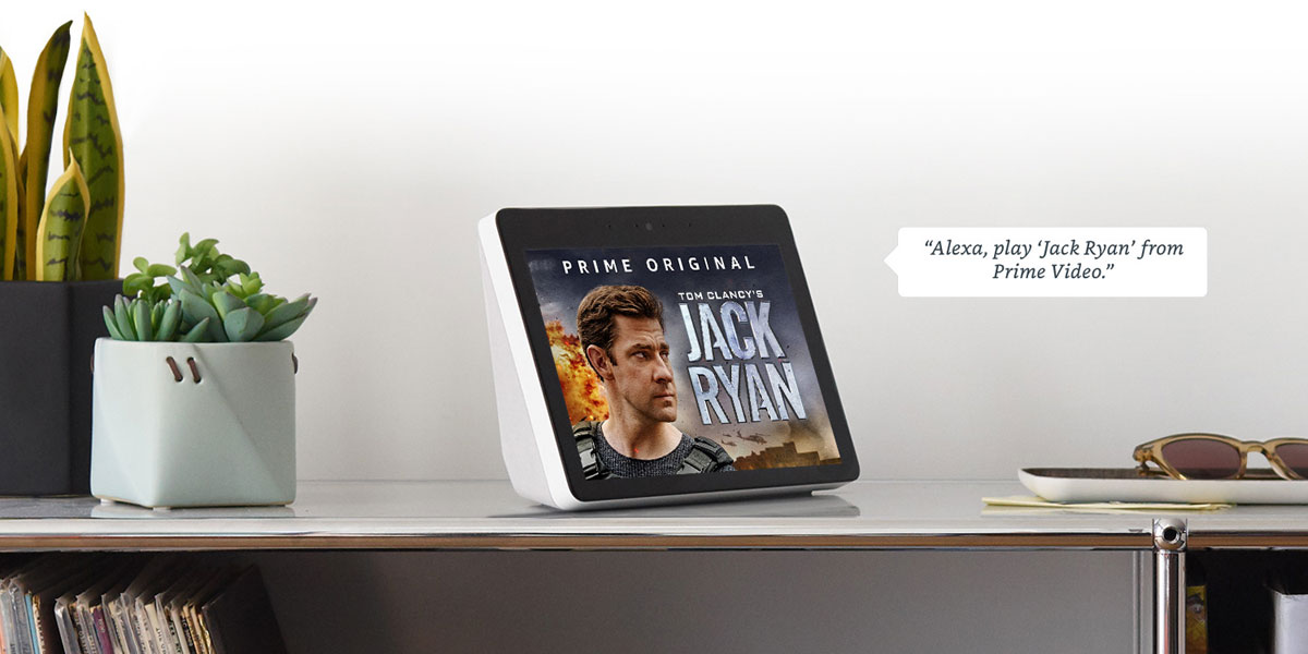   In the center of the picture is an Echo Show on a desk, with potted plants and glasses next to it. A dialog box to the right contains texts reading as “Alexa, play ‘Jack Ryan’ from Prime Video”. And the Echo Show’s screen shows a poster of Tom Clancy’s Jack Ryan, with texts on upper part of the screen reading as “Prime Original” 